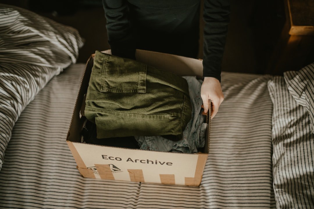 A person packs folded clothing into a cardboard box on a bed.